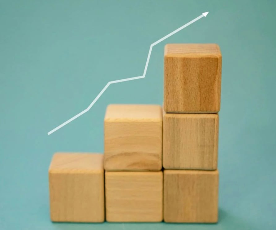 Photo of wooden building blocks stacked on top of each other against a blue background with a white arrow up and to the right.